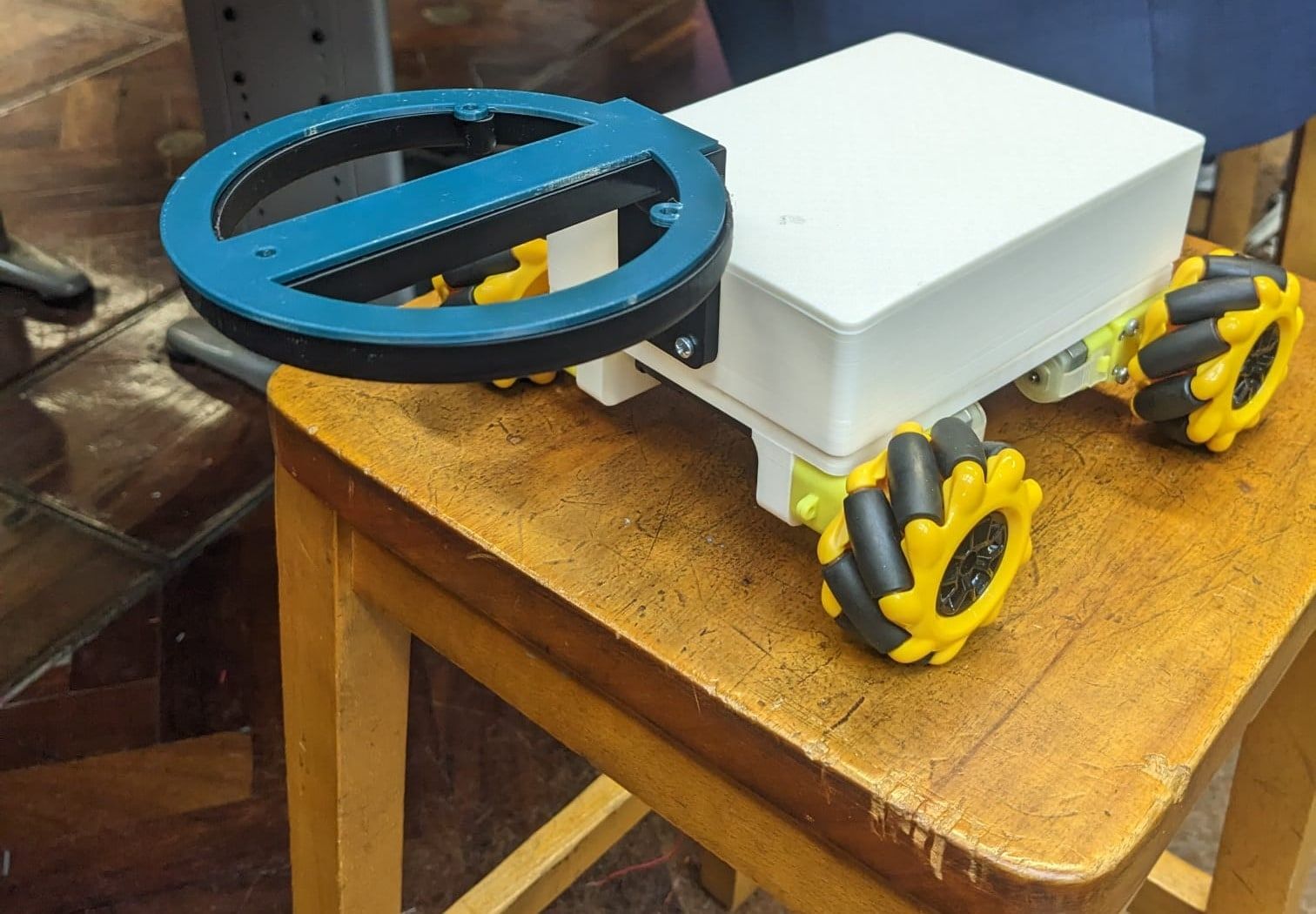 The finished rover on a lab stool: a white 3D-printed enclosure on yellow mecanum wheels, with a blue ring at the front holding the radio antenna coil.